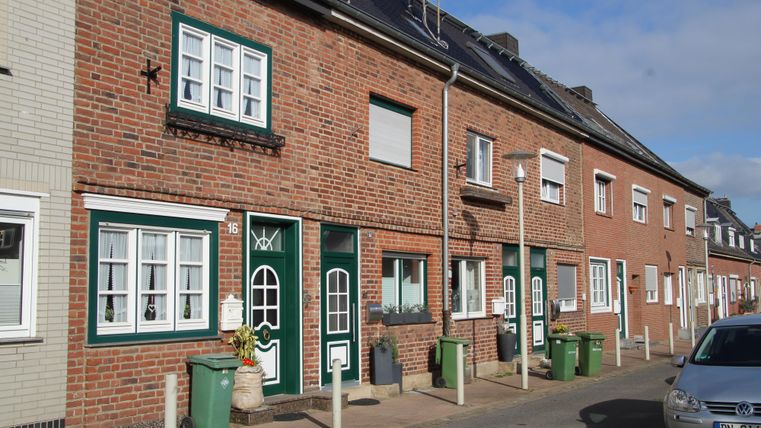 Eine ruhige Straße mit mehreren roten Backsteinhäusern. Die Häuser haben grüne Türen und Fensterrahmen sowie Mülltonnen vor den Eingängen.