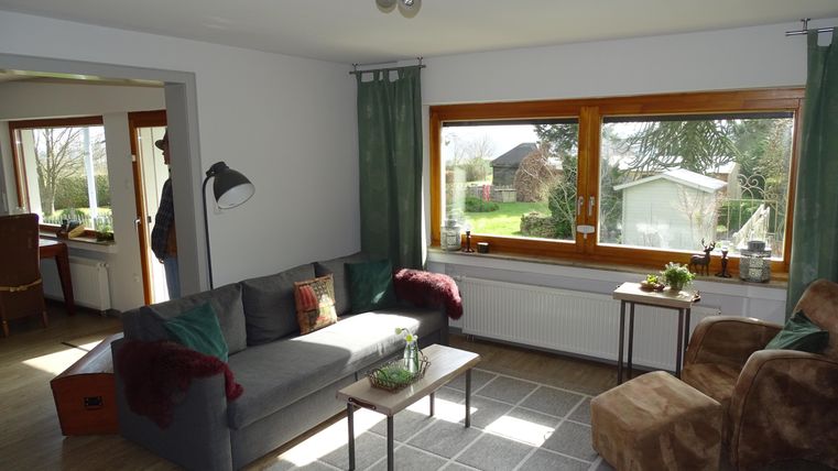 Ein gemütliches Wohnzimmer mit einem grauen Sofa und einem beigen Sessel. Große Fenster bieten einen Blick auf den Garten.