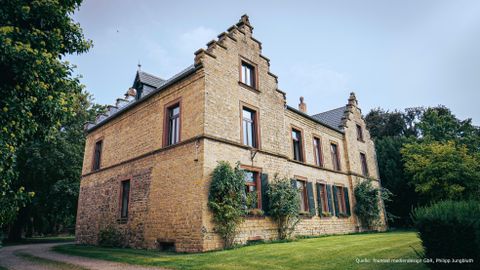 Ein historisches Steinhaus mit Spitzdächern und nostalgischem Charme. Umgeben von einem grünen Garten und Bäumen.