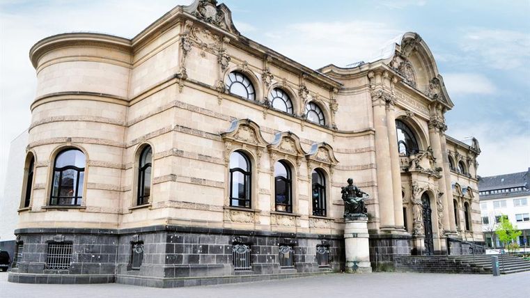 Ein historisches Gebäude mit einer eindrucksvollen Fassade und großen Fenstern. Die Architektur zeigt klassische Elemente und einen schönen Garten davor.