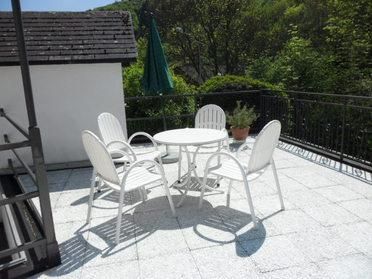 Ein gemütlicher Balkon mit einem runden Tisch und vier weißen Stühlen. Im Hintergrund ist ein grüner Sonnenschirm und viel grünes Laub zu sehen.