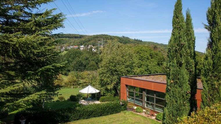 Ein modernes Gebäude umgeben von Bäumen und grüner Landschaft. Im Hintergrund sind sanfte Hügel und ein blauer Himmel zu sehen.