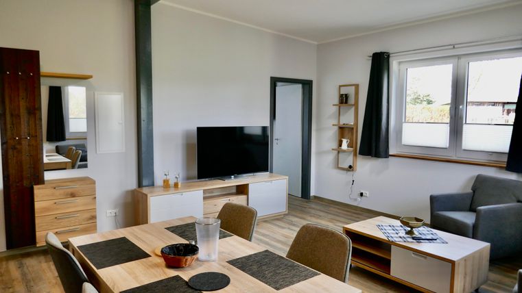 Ein modernes Wohnzimmer mit einem großen Fernseher, einem Esstisch und einer gemütlichen Couch. Die Wände sind hell und die Fenster bieten viel Tageslicht.