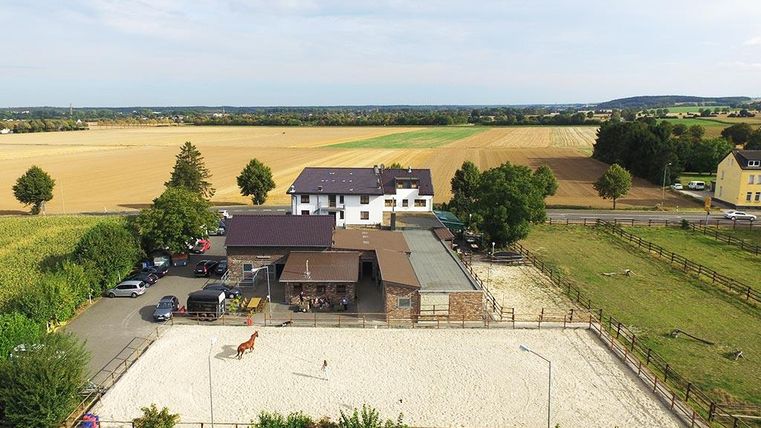 Ein Reiterhof in ländlicher Umgebung mit einem großen Pferdehof. Die Landschaft ist von Feldern und Bäumen umgeben.