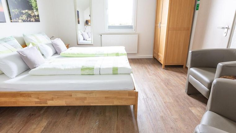 Ein helles Zimmer mit zwei Betten und Holzfußboden. Es gibt einen Stuhl und einen Kleiderschrank, sowie einen Spiegel und ein Fenster.
