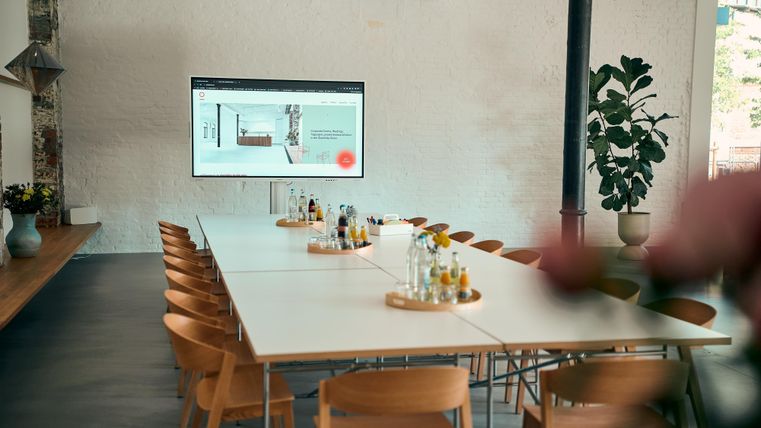 Ein moderner Konferenzraum mit einem langen Tisch und Holzstühlen. An der Wand hängt ein Bildschirm mit Präsentationsinhalten.
