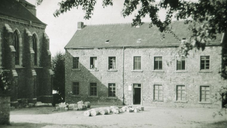 Ein altes Steingebäude mit vielen Fenstern steht auf einem Hof. Einige Säcke liegen auf dem Boden und Bäume werfen Schatten auf die Szene.