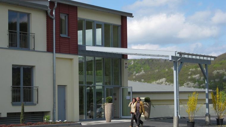 Ein modernes Gebäude mit großen Fenstern und einem überdachten Eingangsbereich. Im Vordergrund sind einige Pflanzen und zwei Personen zu sehen, die sich unterhalten.