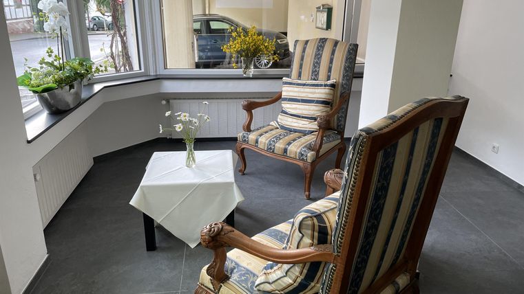 Ein gemütlicher Raum mit zwei Stühlen und einem kleinen Tisch. Am Fenster stehen Pflanzen, und der Raum hat viel Tageslicht.
