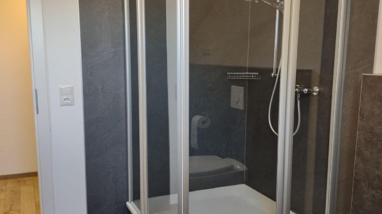 Eine moderne Duschkabine mit klaren Glaswänden steht in einem hellen Badezimmer. Der Boden ist mit Holzplatten ausgelegt, und es gibt eine Tür, die zu einem anderen Raum führt.