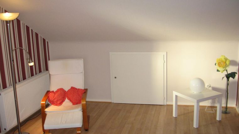 Ein gemütlicher Raum mit einem weißen Sessel und einem kleinen Tisch. An der Wand befinden sich gestreifte Tapeten und eine Lampe sorgt für Licht.