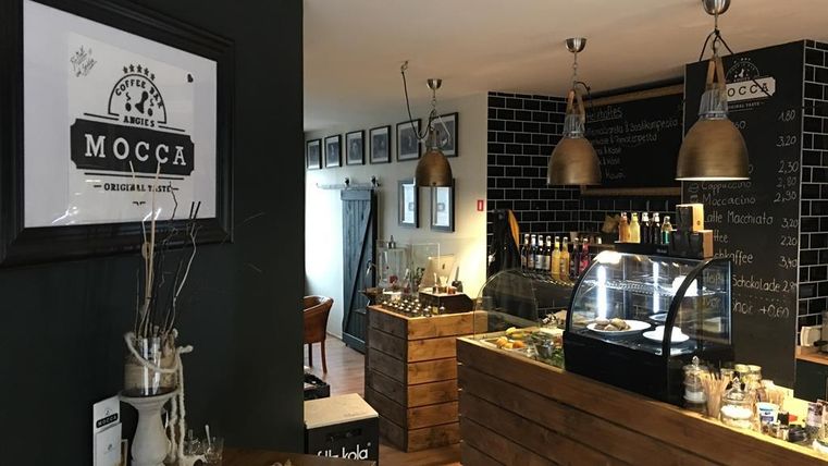Ein gemütliches Café mit Holzregalen und einer Glasvitrine. An der Wand hängen Bilder, und es gibt eine einladende Atmosphäre.