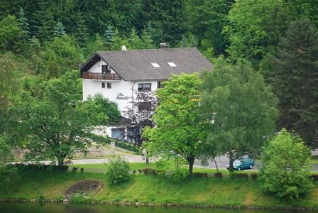 Ein ruhiges Wohnhaus umgeben von grünen Bäumen. Im Vordergrund fließt ein kleiner Weg entlang eines Gewässers.