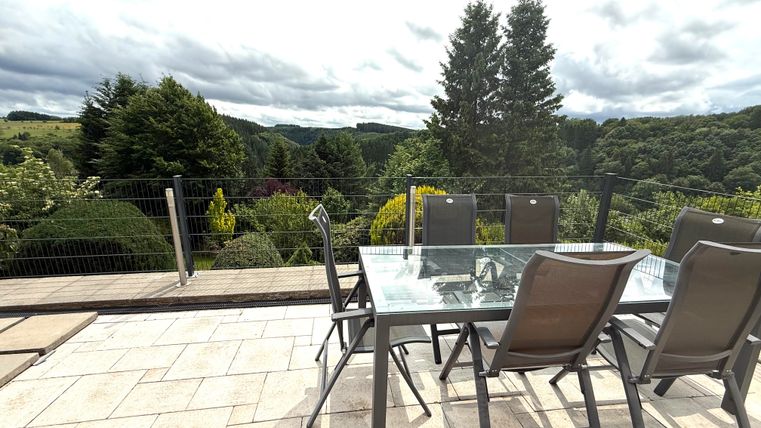 Ein moderner Essbereich im Freien mit einem Tisch und Stühlen auf einer Terrasse. Im Hintergrund sind grüne Hügel und Bäume zu sehen, unter einem bewölkten Himmel.
