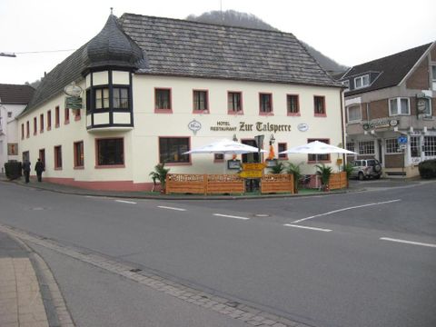 Ein charmantes Restaurant und Hotel namens "Zur Talsperre" an einer Straßenecke. Es hat eine einladende Terrasse mit Sonnenschirmen und ist von einer ruhigen Straße umgeben.