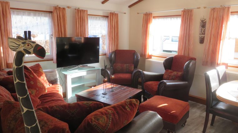 Ein gemütliches Wohnzimmer mit komfortablen Möbeln und einem Fernseher. Große Fenster lassen viel Tageslicht herein und verleihen dem Raum eine einladende Atmosphäre.