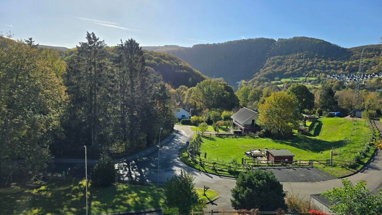 Eine malerische Landschaft mit grünen Wiesen und Bäumen. Im Hintergrund sind sanfte Hügel und ein blauer Himmel sichtbar.