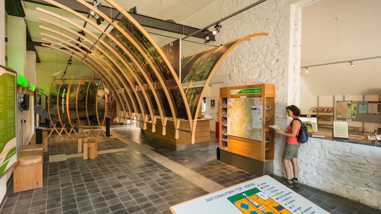 Ein moderner Eingang zu einem Informationszentrum mit Holzstrukturen. Im Vordergrund sind Besucher am Informationsstand zu sehen.