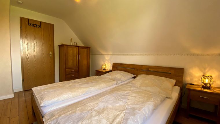 Ein gemütliches Schlafzimmer mit einem Doppelbett, Holzfußboden und warmem Licht. An der Wand steht ein Holzschrank und ein Nachttisch mit einer Lampe.