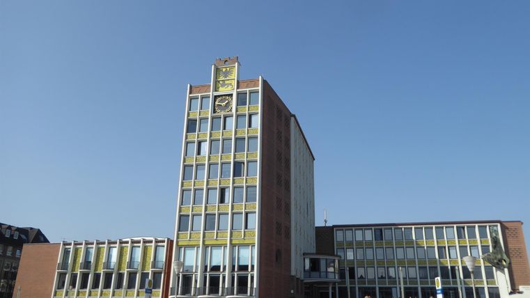 Ein modernes Gebäude mit einer markanten Fassade und einem hohen Turm. Menschen stehen davor und genießen den sonnigen Tag.