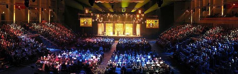 Ein großes Publikum in einer hell erleuchteten Halle. Auf der Bühne finden eine Veranstaltung oder eine Aufführung statt.