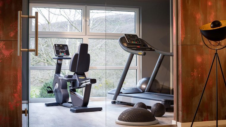 Ein modernes Fitnessstudio mit einem Laufband und einem Liegefahrrad. Große Fenster bieten einen Blick ins Freie und viel natürliches Licht.