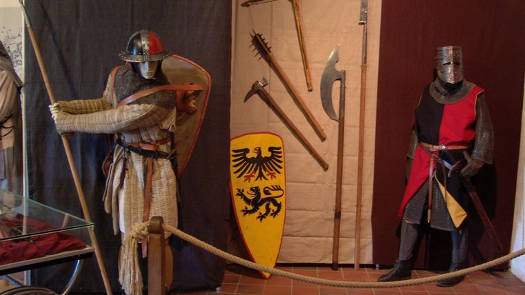 Ein mittelalterliches Museum mit zwei Figuren in Rüstung. Im Hintergrund sind verschiedene Waffen und ein Schild zu sehen.