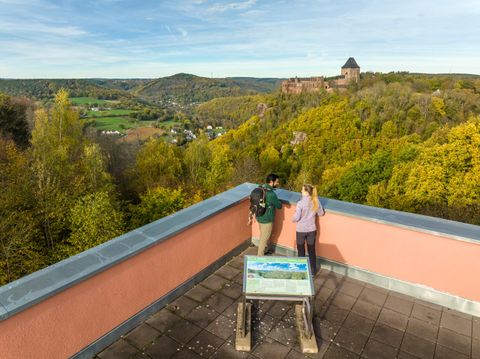 Eine Aussicht von einem Aussichtspunkt mit zwei Personen, die auf die umliegende Landschaft blicken. Im Hintergrund ist eine Burgruine auf einem Hügel zu sehen, umgeben von bunten Bäumen.