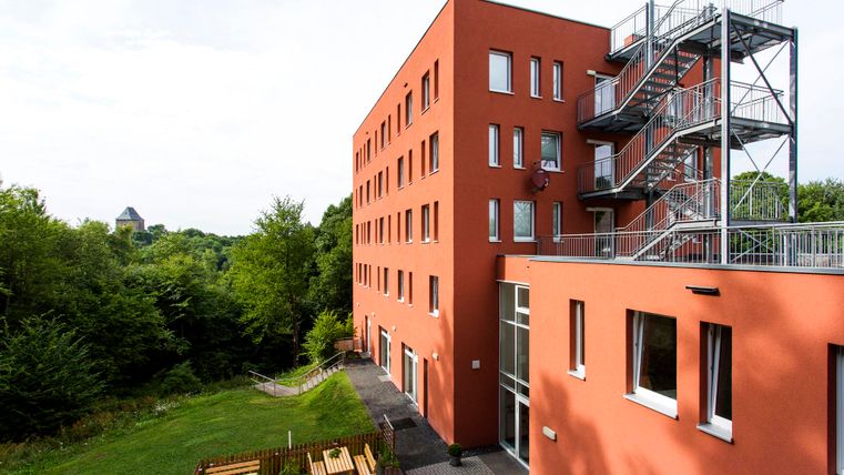 Ein modernes, rotes Gebäude mit einem Außenausgang und einer Feuerleiter. Im Hintergrund sind Bäume und ein Hügellandschaft zu sehen.