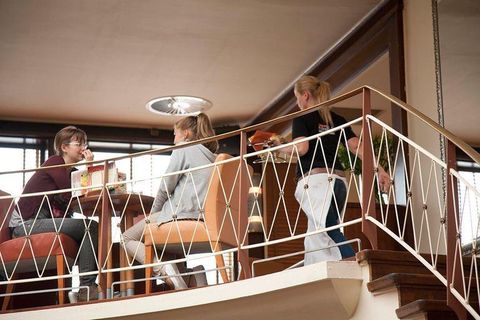 Drei Frauen sitzen in einem Café und unterhalten sich. Eine Kellnerin serviert Essen an einem Tisch.