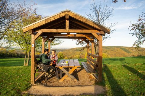 Eine gemütliche Holzlaube auf einer Wiese mit einem wunderschönen Ausblick. Zwei Personen sitzen an einem Tisch und unterhalten sich in der Natur.