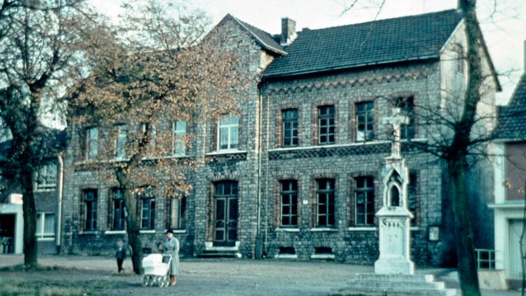 Ein altes Gebäude mit roten Ziegeln und großen Fenstern steht im Vordergrund. Im Hintergrund sind einige Bäume und eine Statue zu sehen.