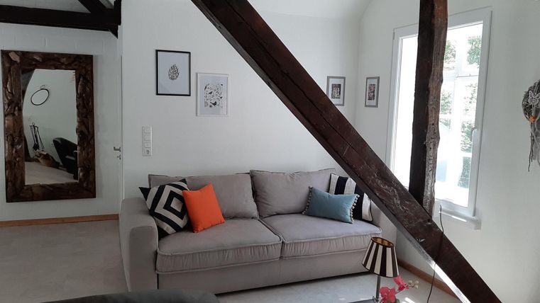 Ein gemütliches Wohnzimmer mit einem grauen Sofa und bunten Kissen. Die Wände sind teilweise mit Holzrahmen dekoriert, und es gibt große Fenster, die viel Licht hereinlassen.