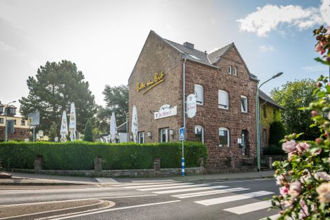 Ein gemütliches Restaurantgebäude an einer Straßenecke. Umgeben von grünen Hecken und Sonnenschirmen sitzen Gäste im Freien.