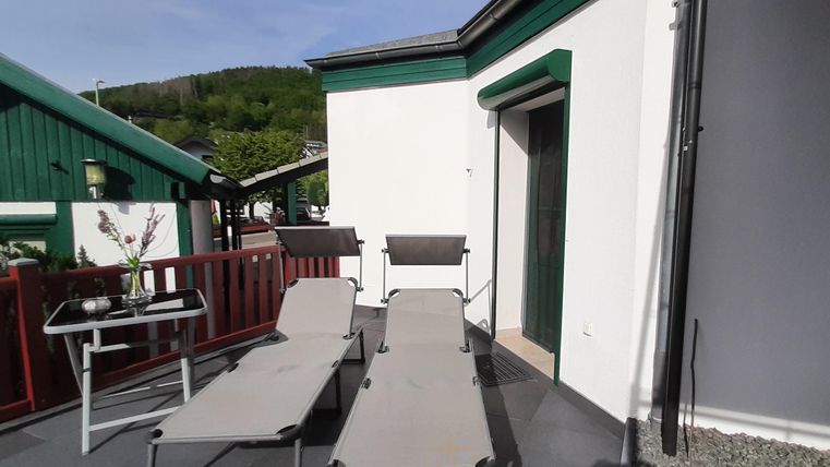 Eine sonnige Terrasse mit zwei Liegen und einem kleinen Tisch. Im Hintergrund sind grüne Gebäude und eine angenehme Landschaft zu sehen.
