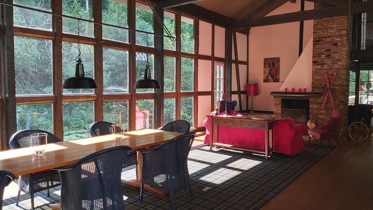 Ein helles Wohnzimmer mit großen Fenstern und viel Tageslicht. Es gibt einen Esstisch, Stühle und einen gemütlichen Bereich mit einem roten Sofa.