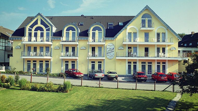 Ein modernes Gebäude mit gelber Fassade und mehreren Balkonen. Vor dem Haus stehen mehrere Autos und eine gepflegte Grünfläche.
