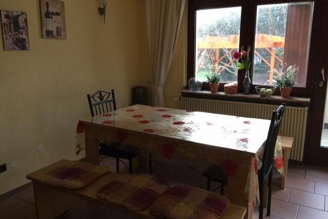 Ein einfacher Essbereich mit einem ovalen Tisch und Stühlen. Große Fenster lassen viel Licht herein, und Blumen dekorieren die Fensterbank.