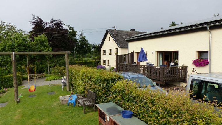 Ein ruhiger Garten mit einer Schaukel und einem Holzdeck. Im Hintergrund steht ein zweigeschossiges Haus mit blühenden Pflanzen.