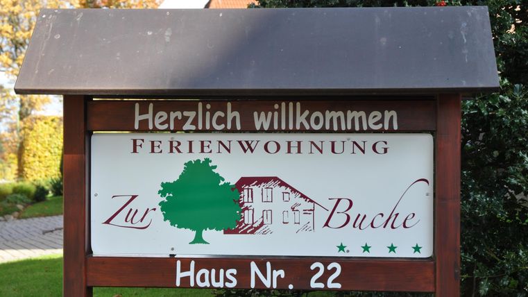 Ein Willkommensschild für eine Ferienwohnung mit dem Namen "Zur Buche". Es zeigt Hausnummer 22 und ist von grünen Pflanzen umgeben.