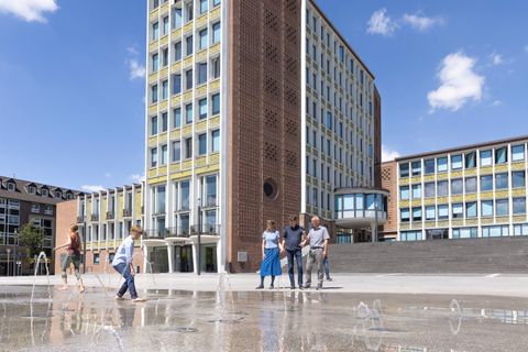 Eine Gruppe von Personen steht vor einem modernen Gebäude und spielt im Spritzwasser. Der Himmel ist blau mit ein paar Wolken.
