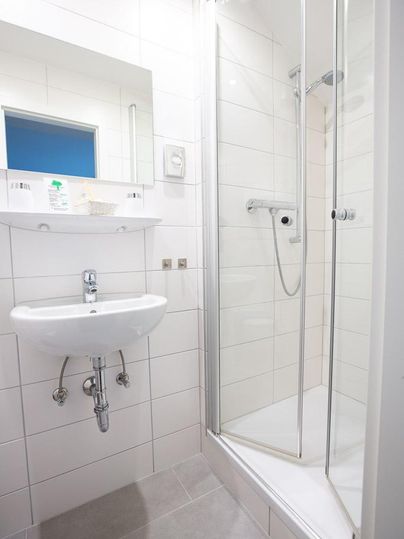 Ein modernes Badezimmer mit einer Dusche und einem Waschbecken. Die Wände sind hell gefliest, und es gibt einen Spiegel über dem Waschbecken.