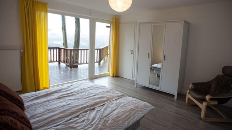 Ein helles Schlafzimmer mit einem großen Bett und einem Ankleiderschrank. Große Fenster mit gelben Vorhängen bieten einen Blick auf die Terrasse.