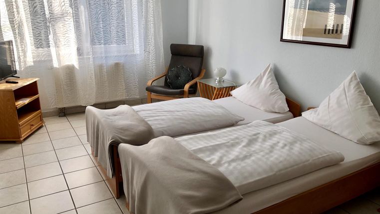 Ein einfaches Schlafzimmer mit zwei Einzelbetten und einem Stuhl. Durch das Fenster fällt natürliches Licht und es gibt einen Fernseher sowie einen kleinen Tisch.