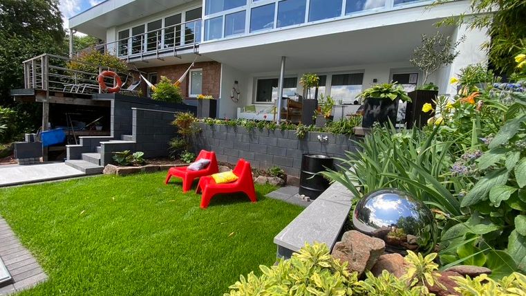 Ein modernes Haus mit großen Fenstern und einem gepflegten Garten. Im Vordergrund stehen rote Stühle und verschiedene Pflanzen, die eine einladende Atmosphäre schaffen.