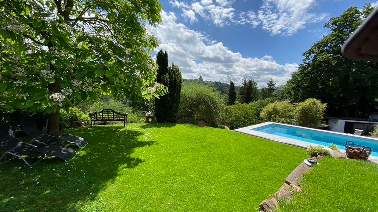 Ein schöner Garten mit grünem Rasen und einem Pool. Der Himmel ist blau mit wenigen Wolken, und es gibt einige Bäume im Hintergrund.