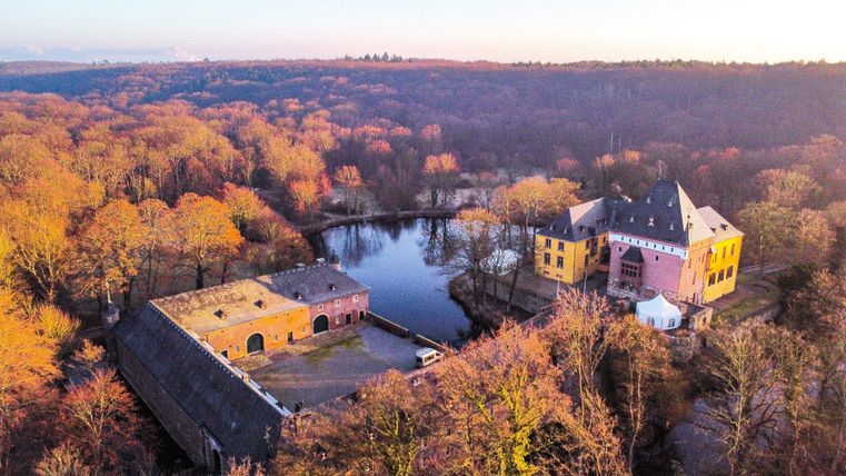 Ein malerisches Schloss am Rand eines klaren Sees, umgeben von buntem Herbstwald. Die Sonne beleuchtet die Szene in warmen Tönen.