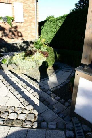 Ein gepflasterter Weg führt durch einen kleinen Garten mit grünen Pflanzen. Im Vordergrund ist der Schatten einer Person sichtbar.