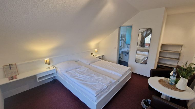 Ein gemütliches Schlafzimmer mit einem Doppelbett und modernen Möbeln. Im Hintergrund sind ein Badezimmer und ein kleiner Wohnbereich sichtbar.