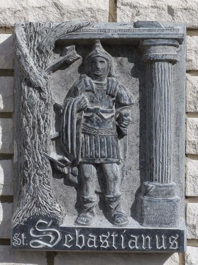 Eine Steinreliefdarstellung von St. Sebastianus in römischer Rüstung. Neben ihm steht ein Baum und eine Säule, mit dem Namen „St. Sebastianus“ unten.